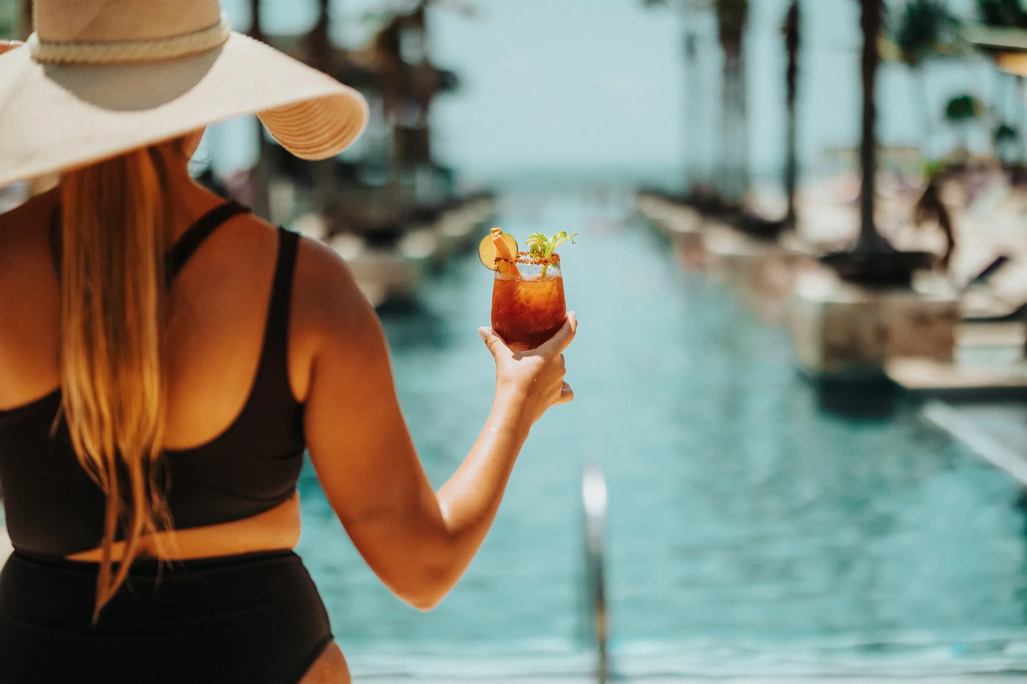 A woman holding a handcrafted Bloody Mary cocktail beside the pool at Breathless Riviera Cancun, overlooking the palm-lined resort grounds.